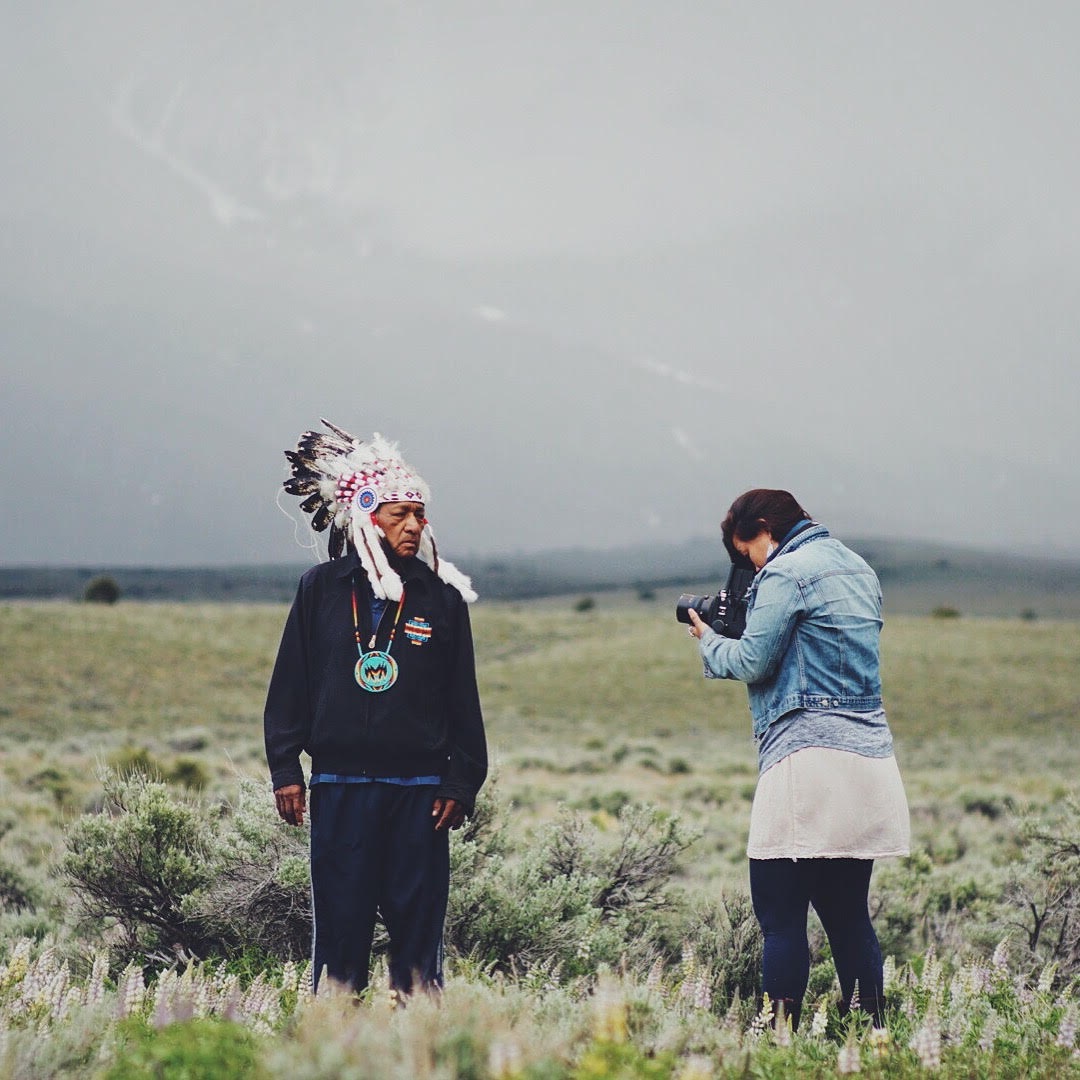 Matika Wilbur outside on the plains taking a picture of an older Native American man with traditional headware