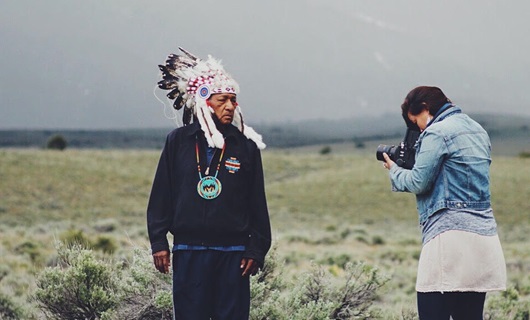 Matika Wilbur outside on the plains taking a picture of an older Native American man with traditional headware