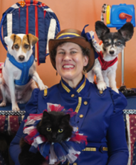 Picture of Sharon Page wearing a blue jacket with gold buttons and trim. She is holding a black cat and two small dogs are behind her shoulders.