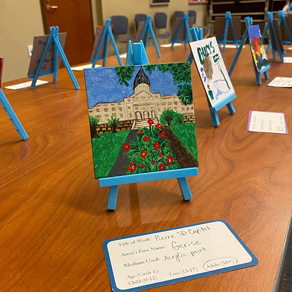 Small painted canvas of a building with roses sitting on a small easel on top of a table. Other small artwork is in the background.,