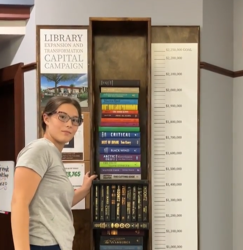 A woman standing next to a bookshelf with books stacked to represent the funds being raised in the library's capital campaign.