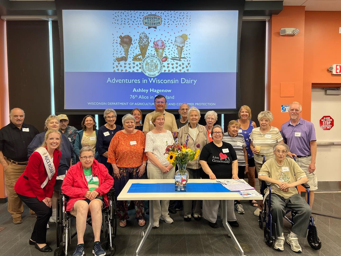 A large group of people posing with Alice in Dairyland in front of a screen that reads "Adventures in Wisconsin Dairy", Ashley Hagenow, 76th Alice in Dairyland."