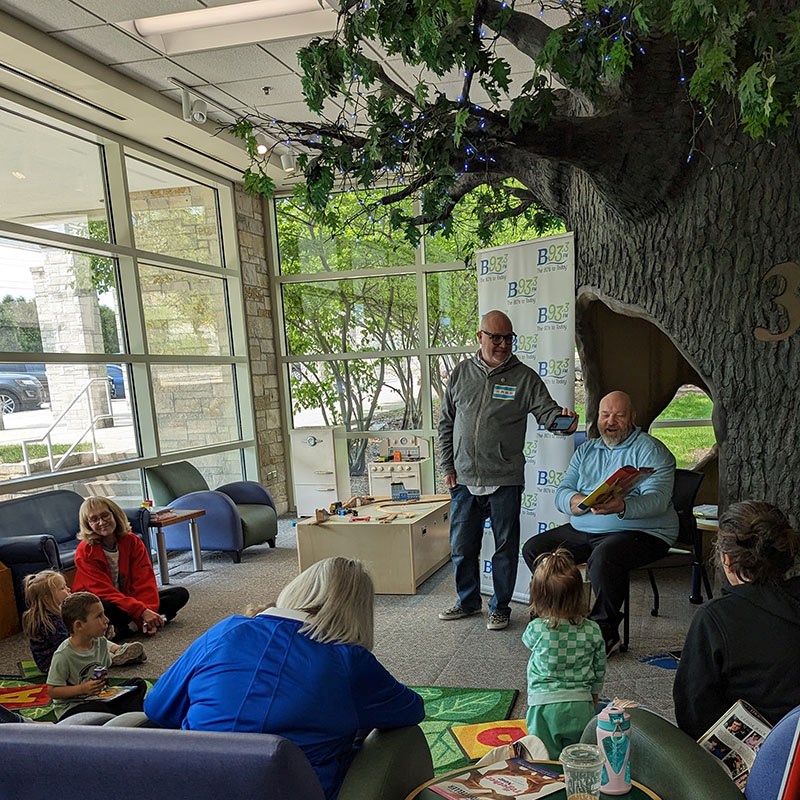 A radio host sits and holds a book reading to a room of children and adults A radio host sits and holds a book reading to a room of children and adults