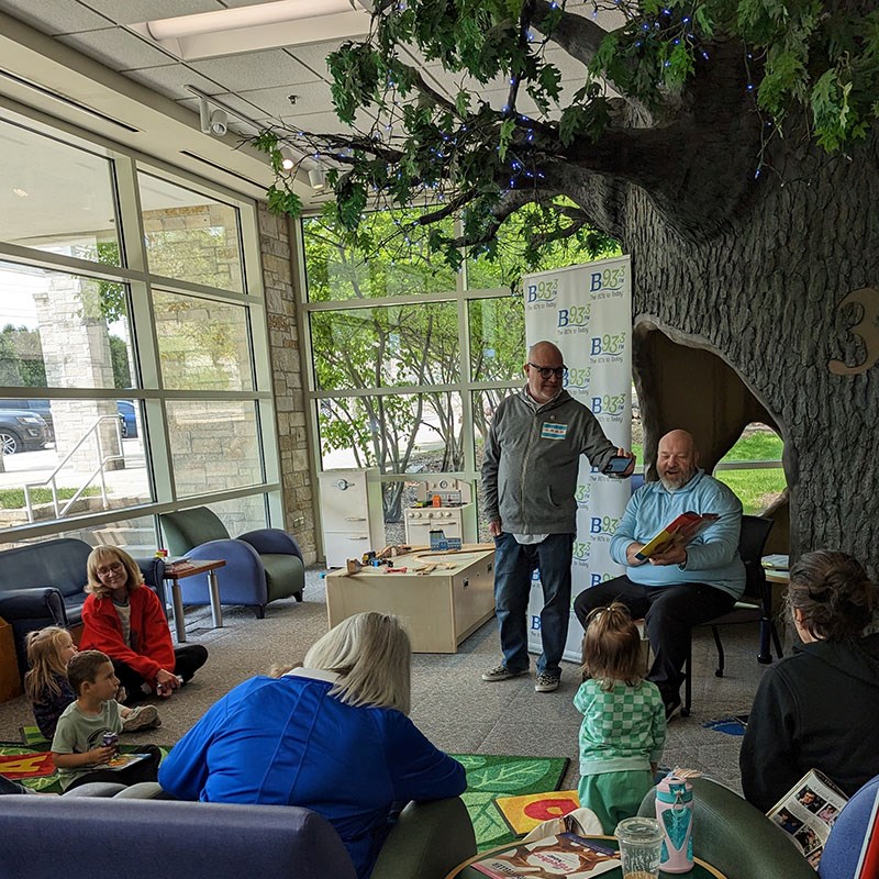 A radio host sits and holds a book reading to a room of children and adults