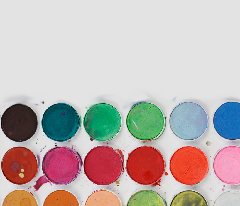 A person's hands painting leaves onto round buttons. There is a palette of green paint and a plant on the tabletop.