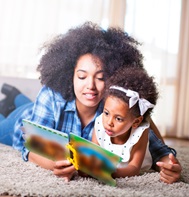 Black woman and baby girl, lying on a rug and reading a book together.