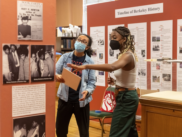 Two female museum curators in front of a display discussing it. Wearing covid-19 masks.