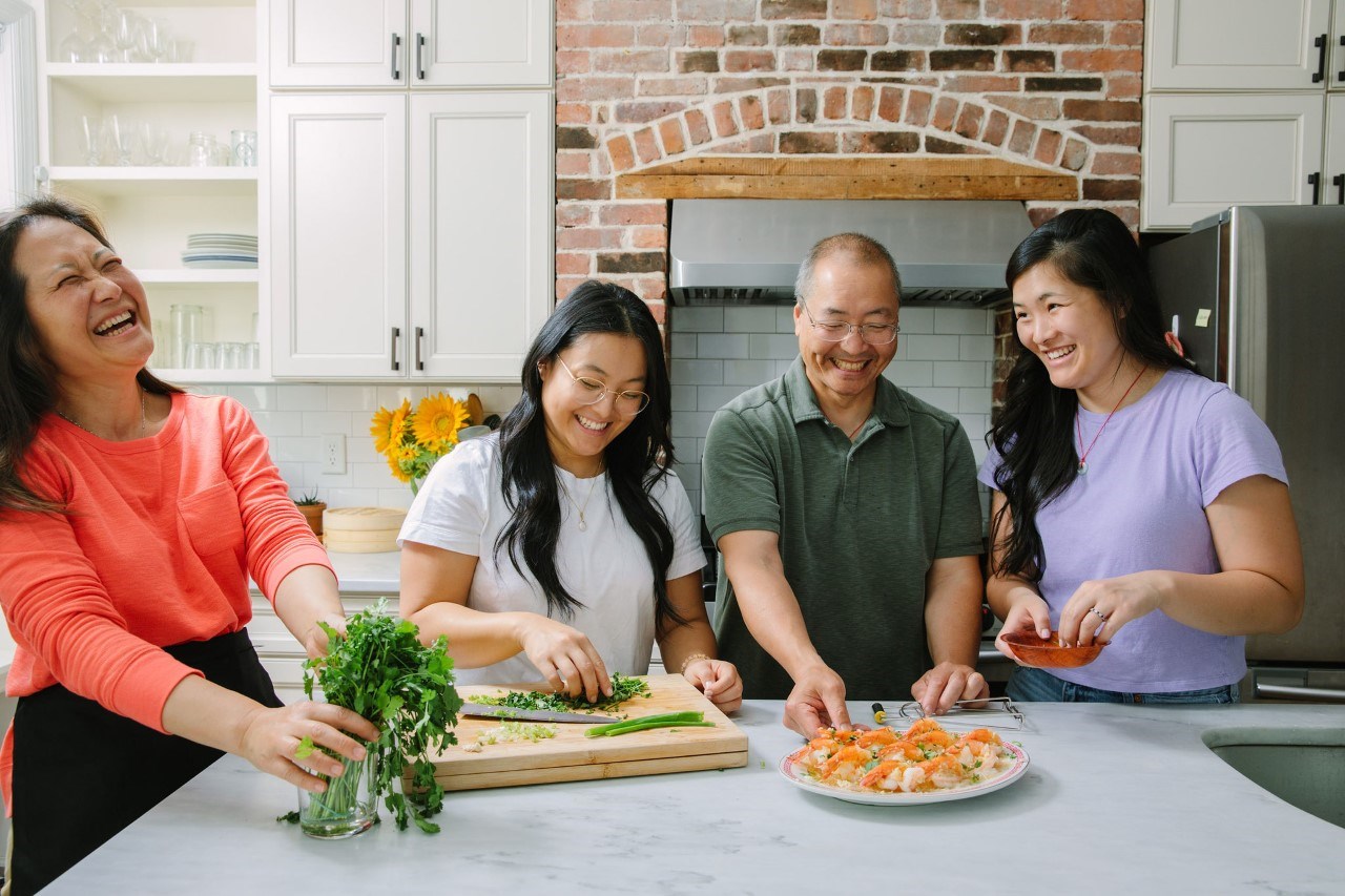 Family of four - mother, father, two adult female children cooking and laughing in kitchen with one another