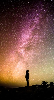 Lone figure in silhouette looks up at dark sky filled with colorful stars