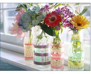 A photo of 4 clear bottles of various shapes and sizes that have been decorated with colorful washi tape and filled with colorful flowers, sitting on a windowsill - courtesy of prettyhandygirl.com
