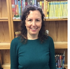 Photo of new youth services librarian Geralyn Schultz standing in front of a bookshelf, smiling