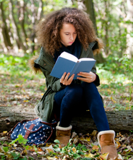 A teenager sits outside on a log, reading a book.