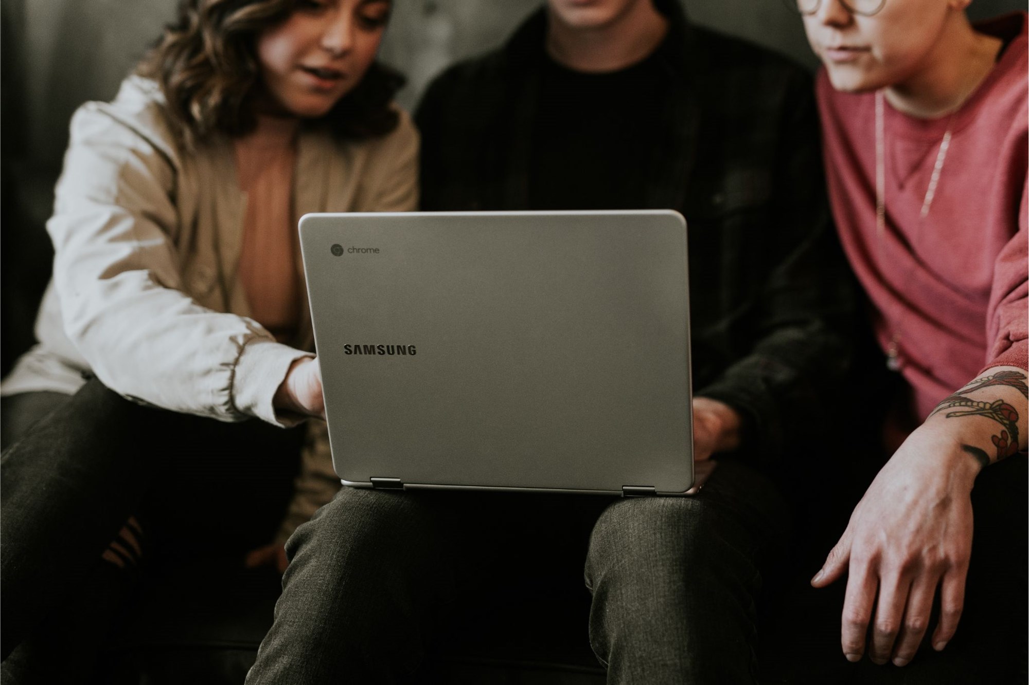 three people sitting and looking at laptop on the middle person's knees three people sitting and looking at laptop on the middle person's knees