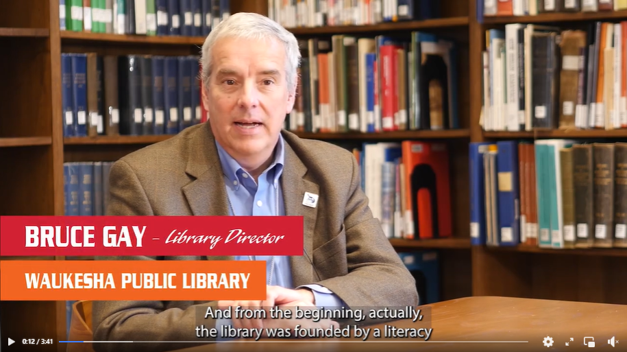 Screenshot of a video from Literacy Services of Wisconsin with Bruce Gay, Waukesha Public Library Director, sitting at a table talking to the camera.