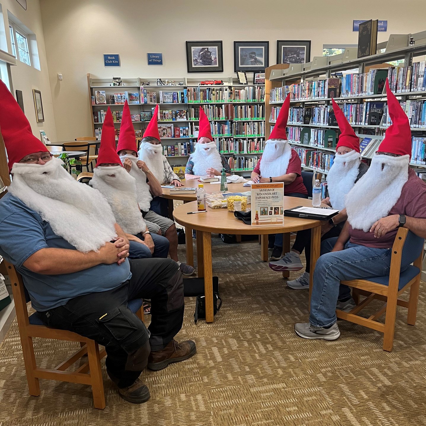 Photo of a group of eight people wearing gnome hats and beards sitting around tables inside the library.