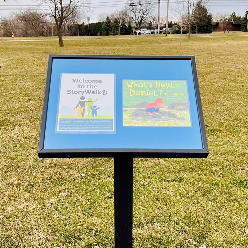 Photo of the first sign of Dwight Foster Public Library's StoryWalk in Ralph Park showing the book, "What's New, Daniel?" by Micha Archer in a park.