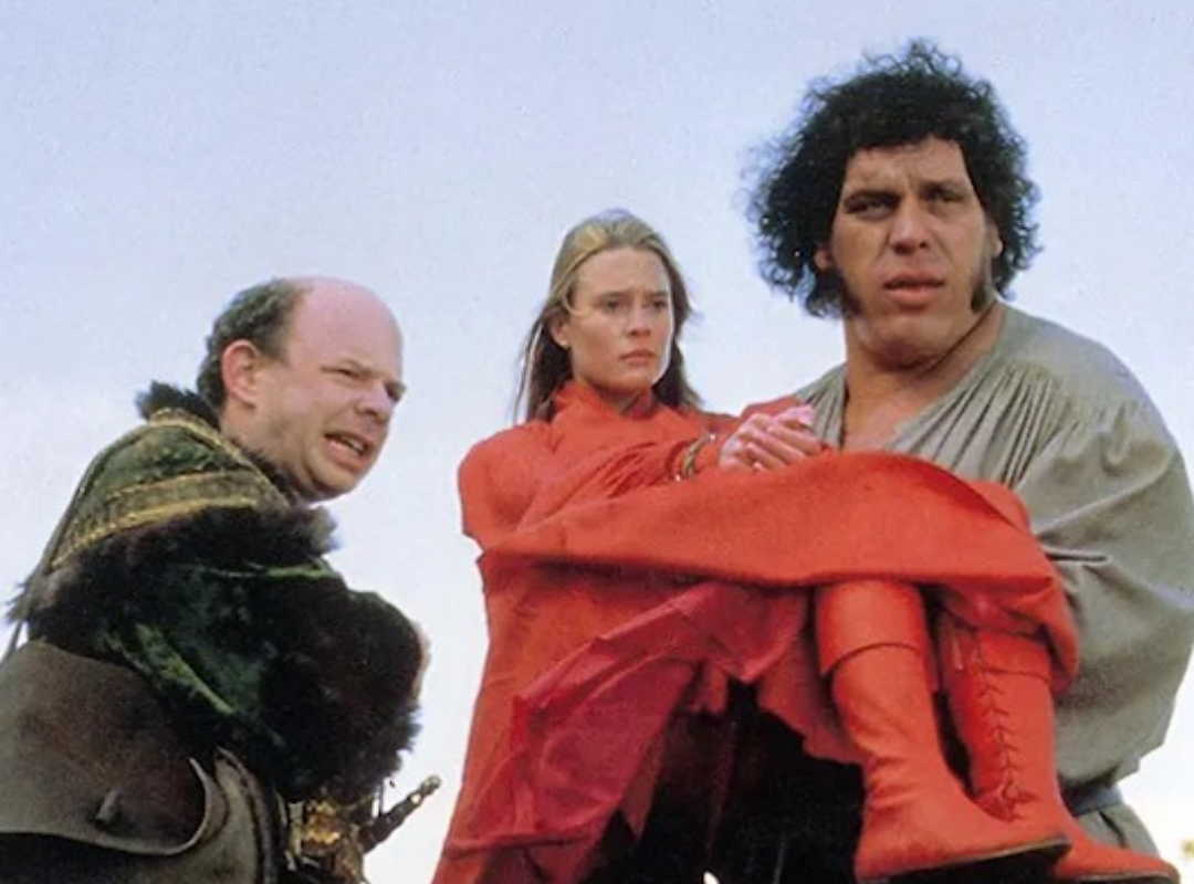 A screenshot from The Princess Bride, depicting a large man carrying a woman dressed entirely in red, alongside a shorter man. All three are looking towards something with an uncertain expression.