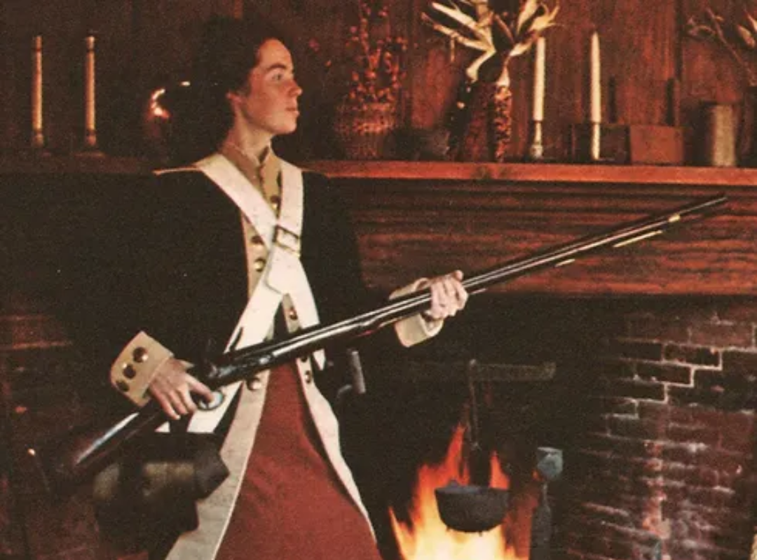 An image showing a woman in historic soldier attire holding a large gun in front of a fireplace.