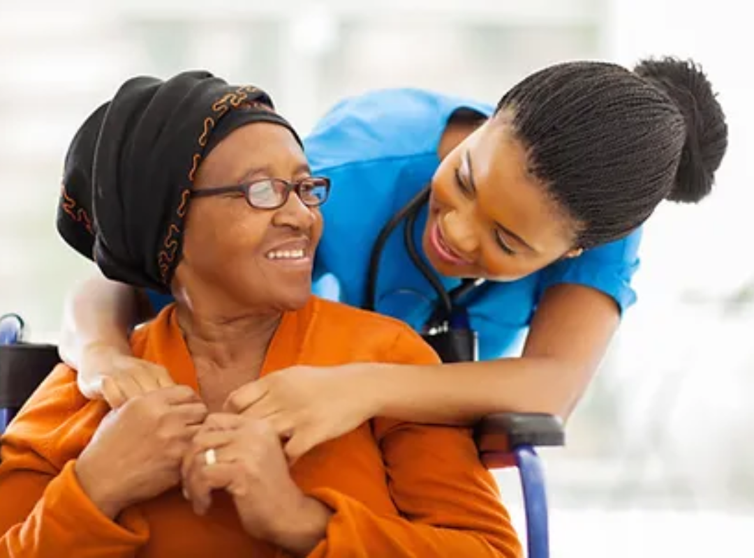A young lady cares for an older lady in a wheelchair, smiling.