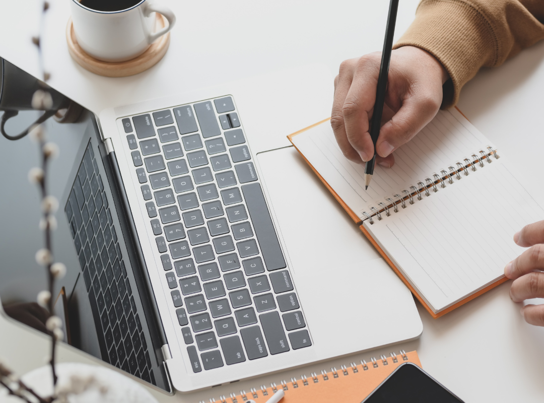 Photograph of a person writing in a notebook alongside a laptop.