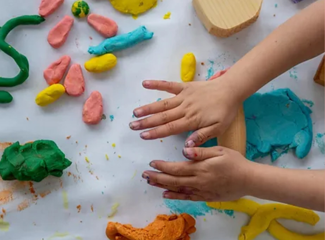 A child's hands rolling colourful pieces of clay into a variety of shapes against a white background