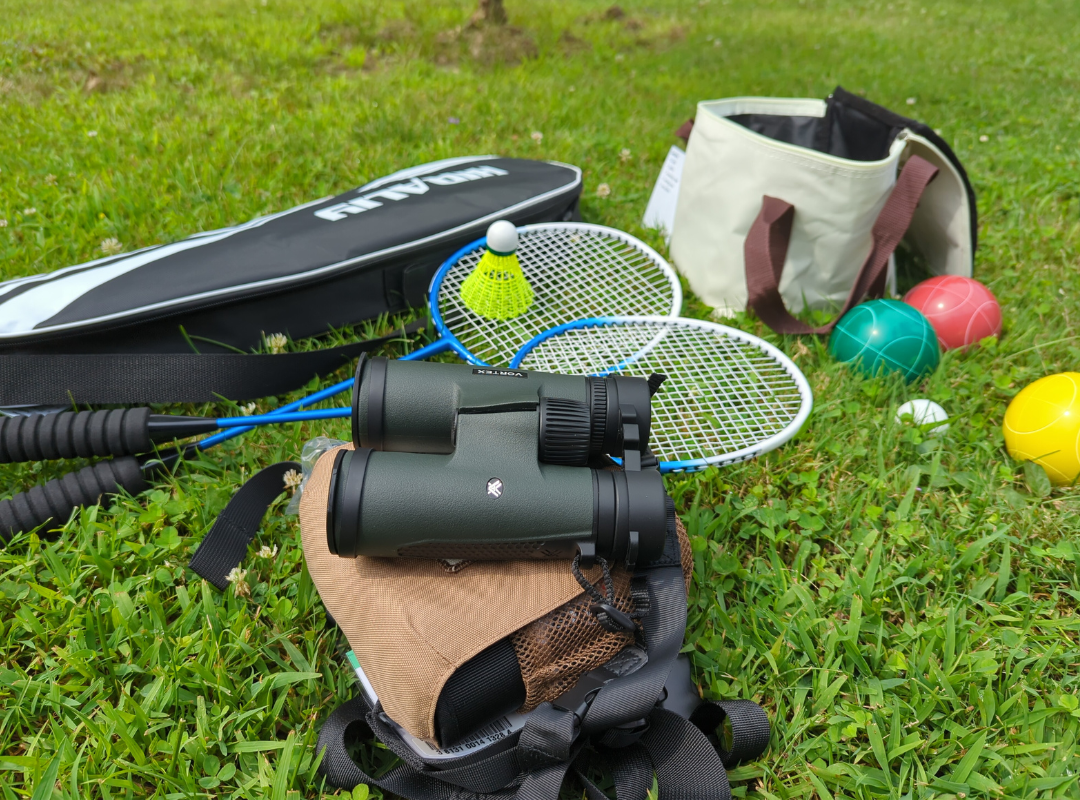 A photograph of badminton rackets with a shuttlecock and case, bocce balls and case, and a pair of binoculars, arranged on grass.