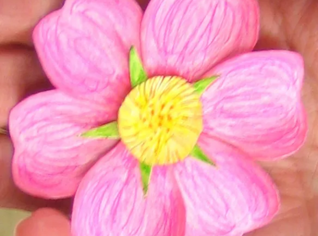 Photograph of a handmade paper flower with pink petals with pleasing, scribbly pencil textures, and a bright yellow centre. 