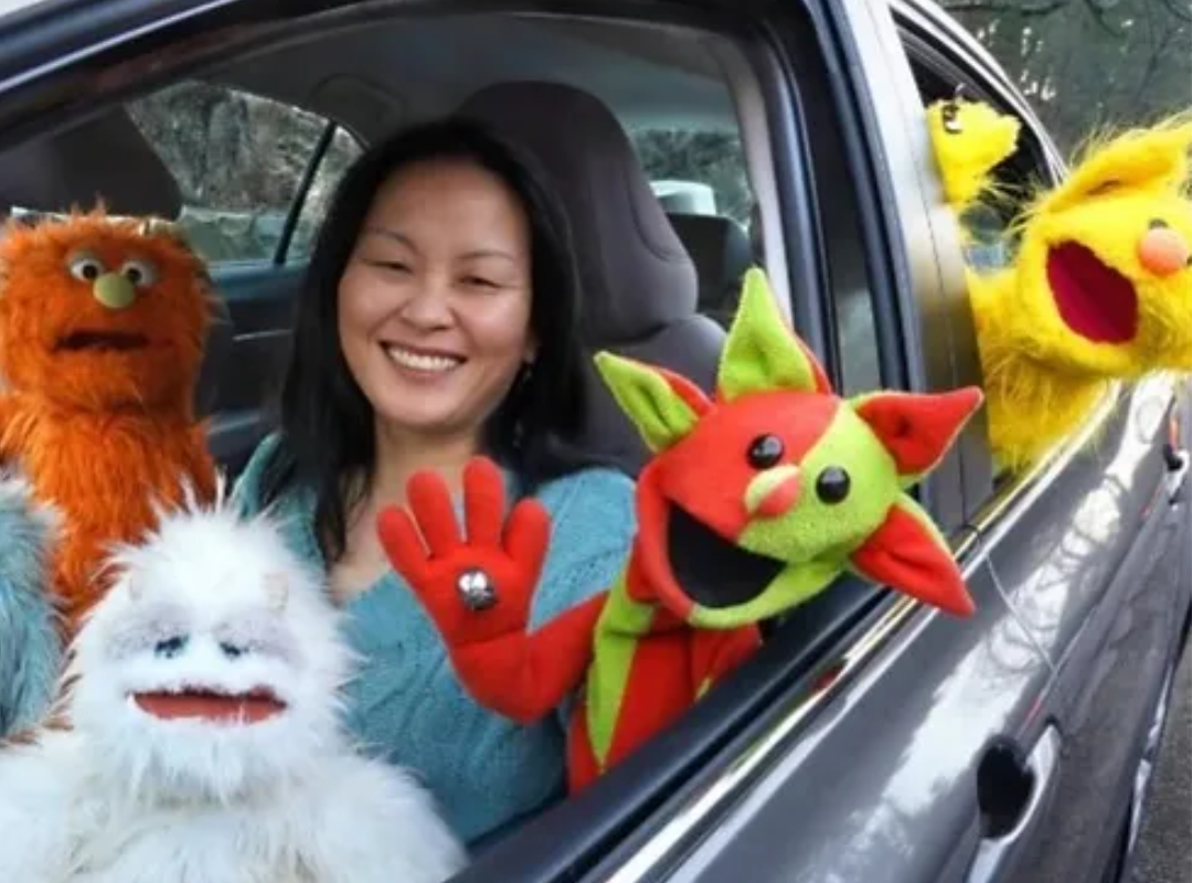 A smiling woman sitting in her car, with four colourful and fuzzy puppets surrounding her and hanging out of the windows. 