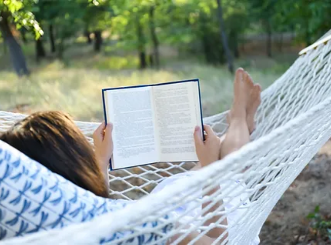 A photo of a person laying in a hammock whilst reading a book. The sunlight illuminates foliage in the background.