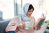 Photo of a smiling woman in a headscarf, seated on a couch, reading a book and holding a mug.