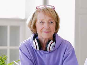 Senior lady with headphones around her neck sitting at a computer desk with laptop