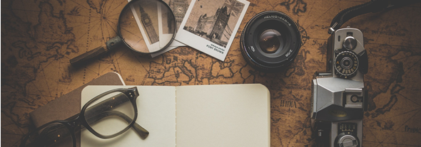 Pair of glasses, magnifying glass, old photographs, camera and notebook on top of an old map