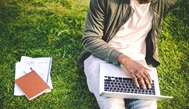 Photo of a man's body, sitting on grass, laptop in his lap, and notebooks beside him in the grass
