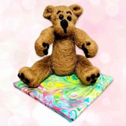 Image links to website calendar.  On a light pink background a brown needle felted bear is sitting on a tie-dye towel.