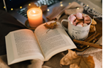 Cozy image of a book, lit candle, soft blanket and hot chocolate with marshmallows.