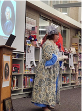 A woman dressed in clothing from the Colonial/Revolutionary era, giving a presentation in the library. The calendar event link this image will take you to will open an external site and in a new tab or window.