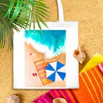Image depicts a canvas bag with a beach design laying on a beach next to a beach towel.