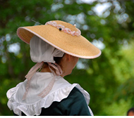 A woman in Revolutionary era clothing showing the back of her collar and her hat.