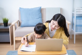 Image depicts a mom helping a child writing notes in a notebook next to a laptop.